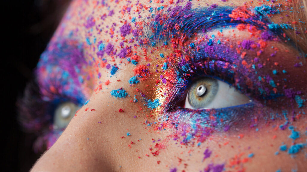 Blaue Augen einer jungen schönen Frau mit einem Lidschattenspray, eine Streuung von Farbe auf ihrem Gesicht, bunter Effekt