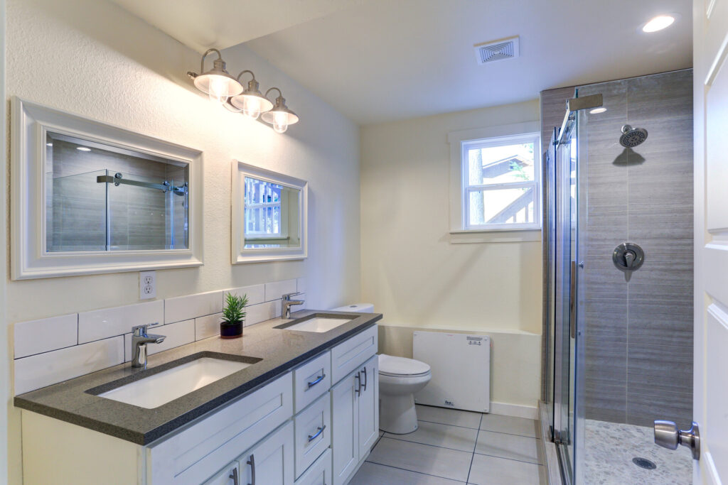 Badezimmer mit schönen Badmöbeln, weißer Badezimmer-Waschtisch mit Granitplatte, Doppelwaschbecken, Doppelwandspiegel mit weißem Holzrahmen und Dusche mit Glastür