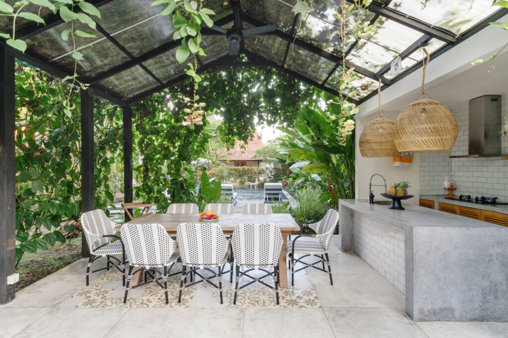 Offene Küche mit Esstisch und Stühlen draußen, vor grünen, frischen Pflanzen im Hintergrund